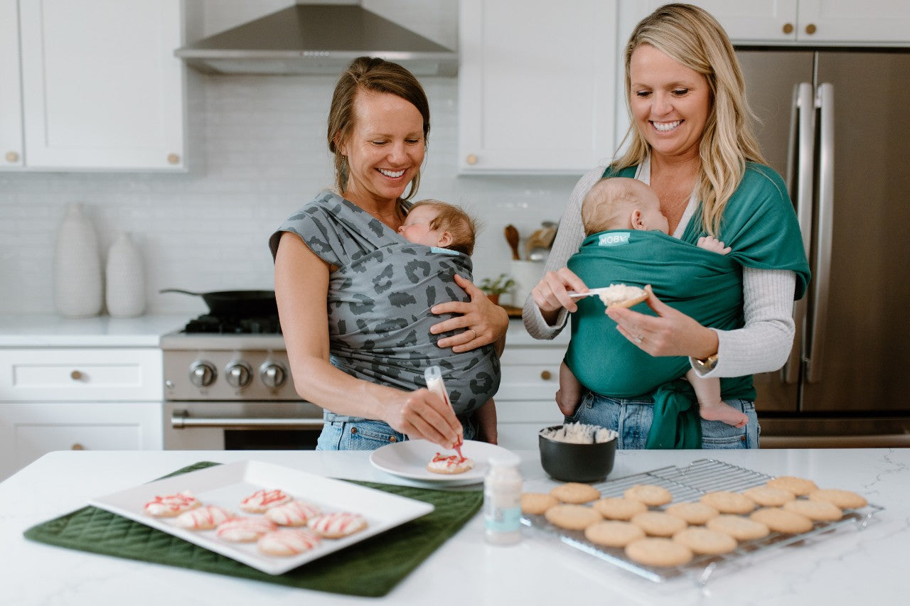 Two mom's wearing their babies in wraps while decorating christmas cookies