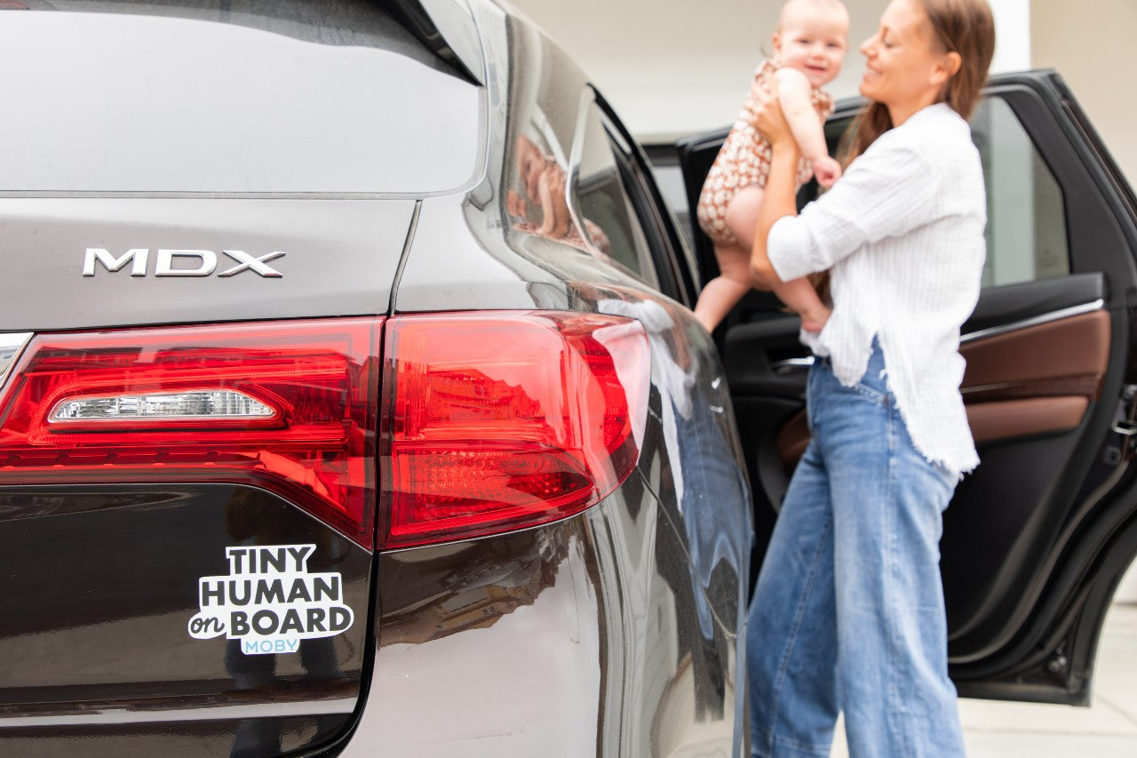 Mom puts baby in car that has a Moby sticker that reads "Tiny Human on Board” on back of car. 