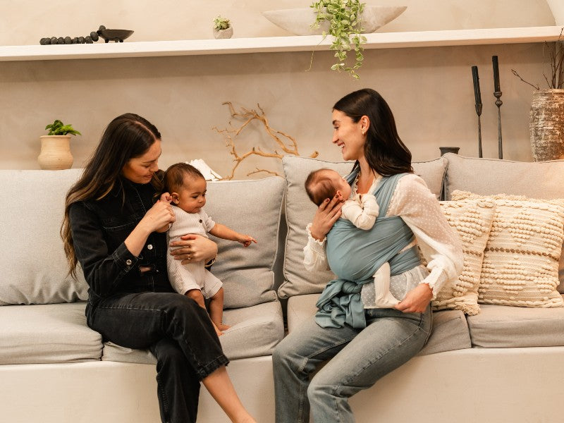 A mom wearing her baby facing inward, sitting on a couch and speaking with another mom holding a baby in her arms 