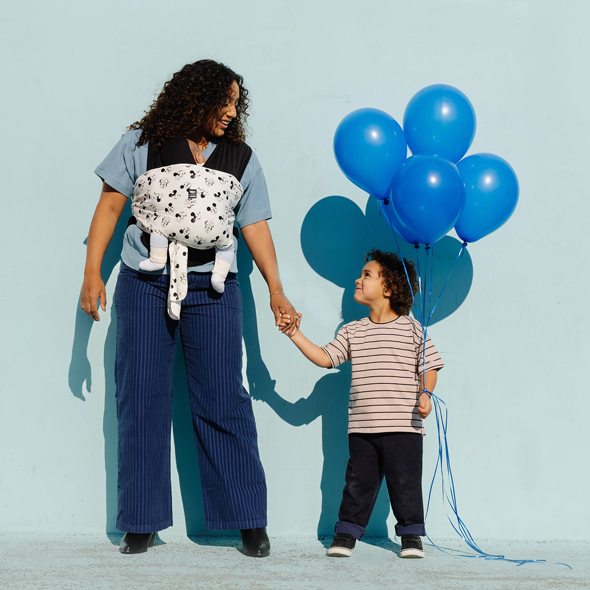 mom wearing baby in Easy-Wrap Carrier in Disney's Mickey Mouse & Minnie Mouse while holding hands with toddler boy who is holding blue balloons in his other hand
