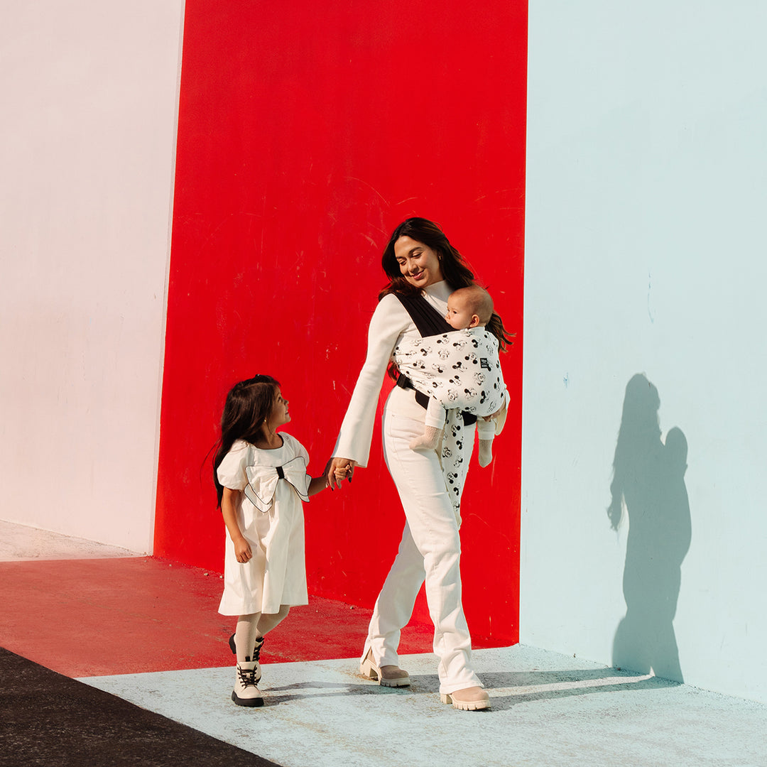 mom wearing baby in Easy-Wrap Carrier in Disney's Mickey Mouse & Minnie Mouse while walking and holding hands with toddler girl