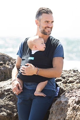 man sitting on rocky beach wearing his baby in a moby wrap