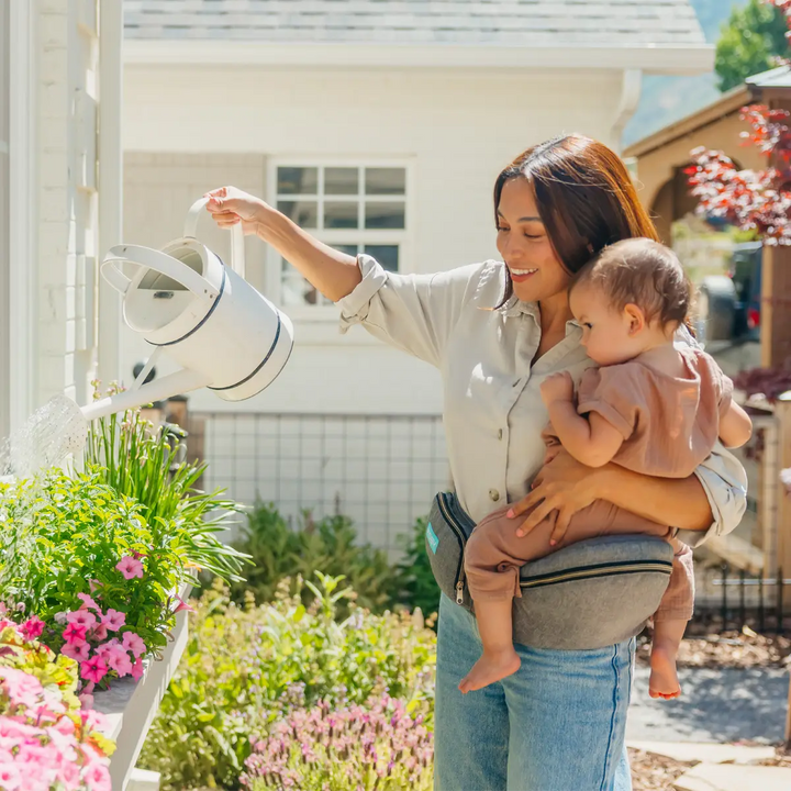 mom wearing baby in hip seat in heather grey while watering the plants and flowers with a watering pot