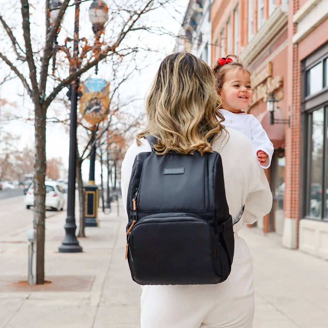 mom wearing the Parent Pack Diaper Bag in Black while holding baby